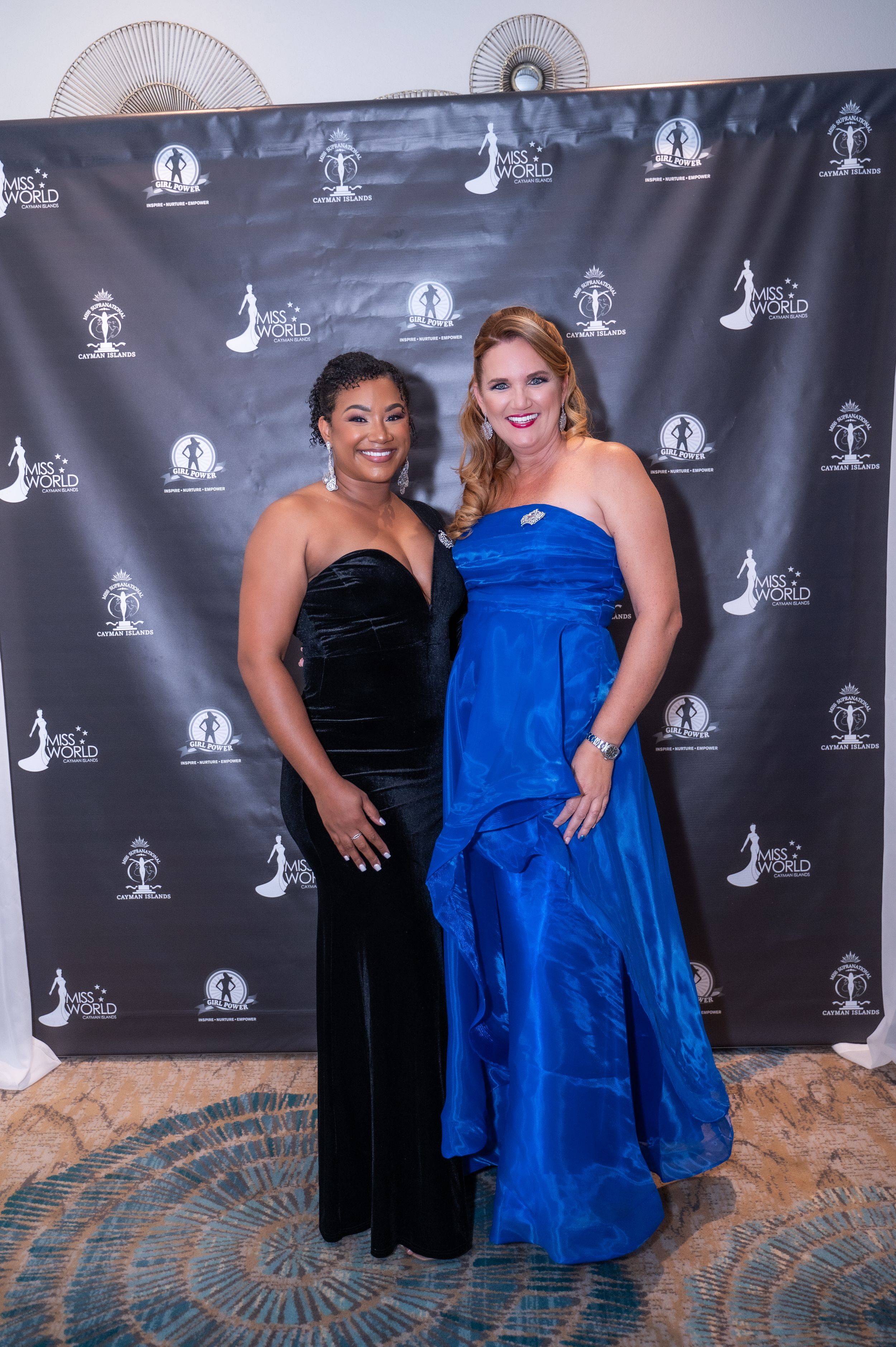 Two elegant women pose at a formal event, one in black velvet gown and one in royal blue dress, before a step-and-repeat backdrop.