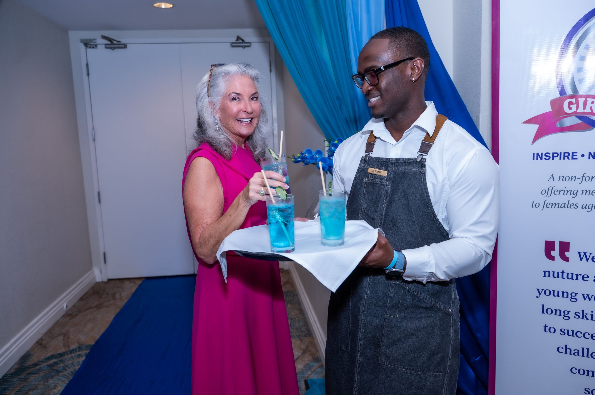 A woman in pink dress accepts a blue cocktail from a server in apron at a formal networking event.