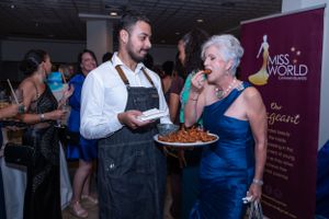 Chef serves food to elegantly dressed woman at Miss World Cayman Islands event in formal venue.