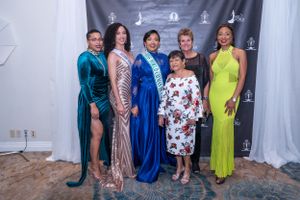 Six women in elegant formal gowns pose together at a pageant event in front of a branded step-and-repeat backdrop.