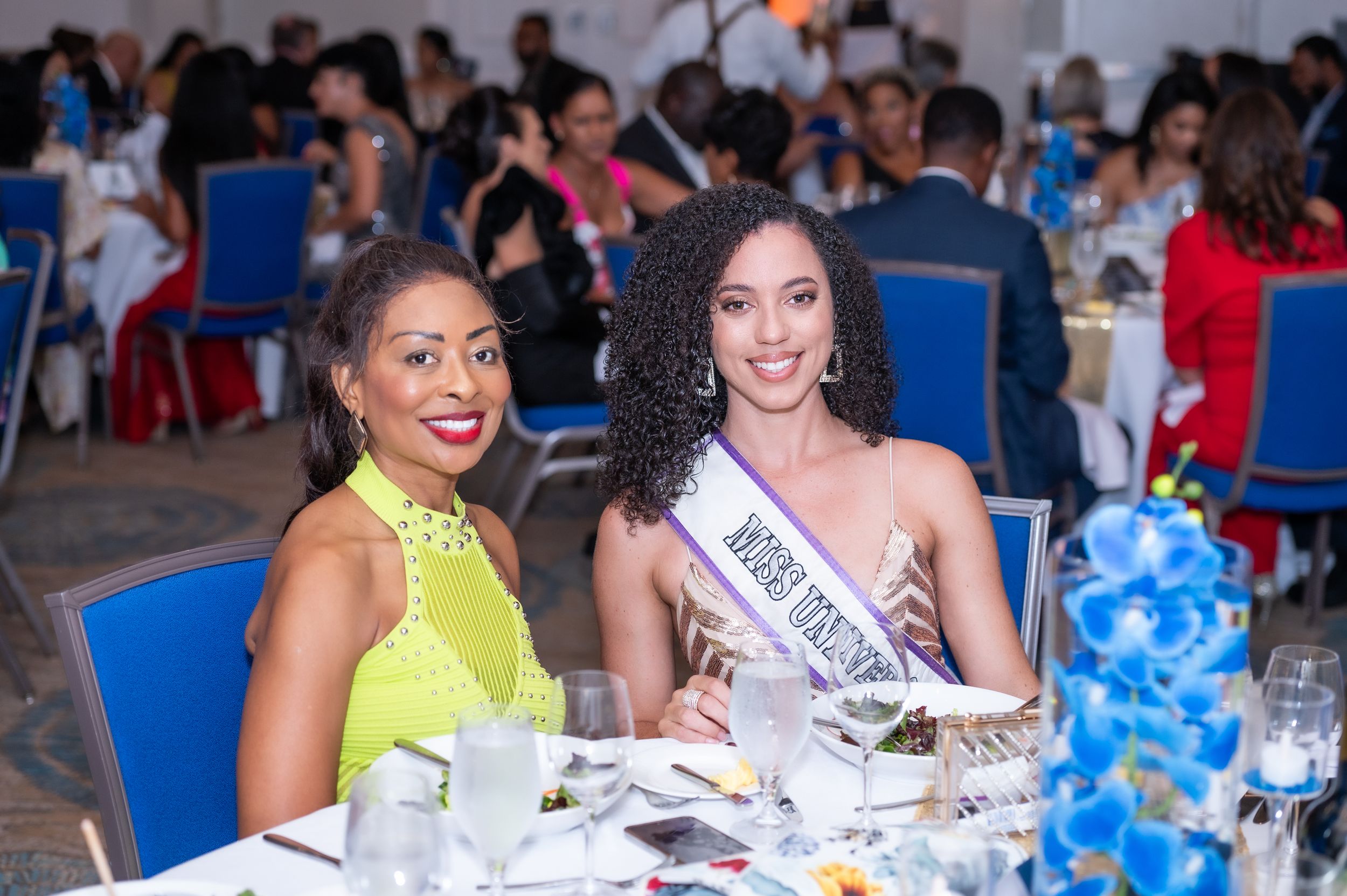 Two elegant women smile at a formal gala dinner, one wearing a beauty pageant sash and blue floral centerpieces nearby.