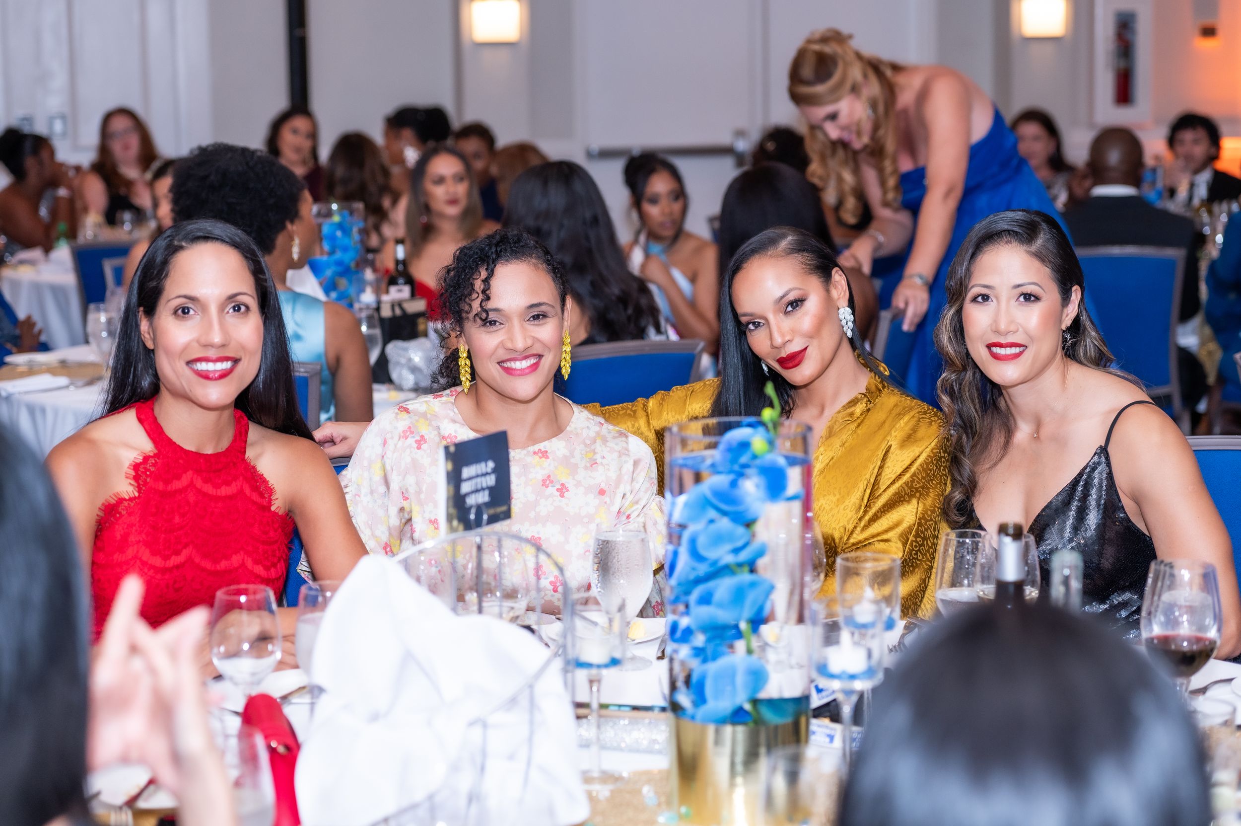 Four elegant women smile at a formal gala dinner, dressed in red, white, gold, and black attire.