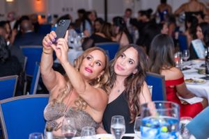 Two women in elegant attire take a selfie at a formal gala dinner event with guests dining in background.