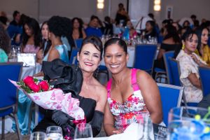 Two smiling women pose together at an elegant banquet event, one holding a bouquet of pink roses.