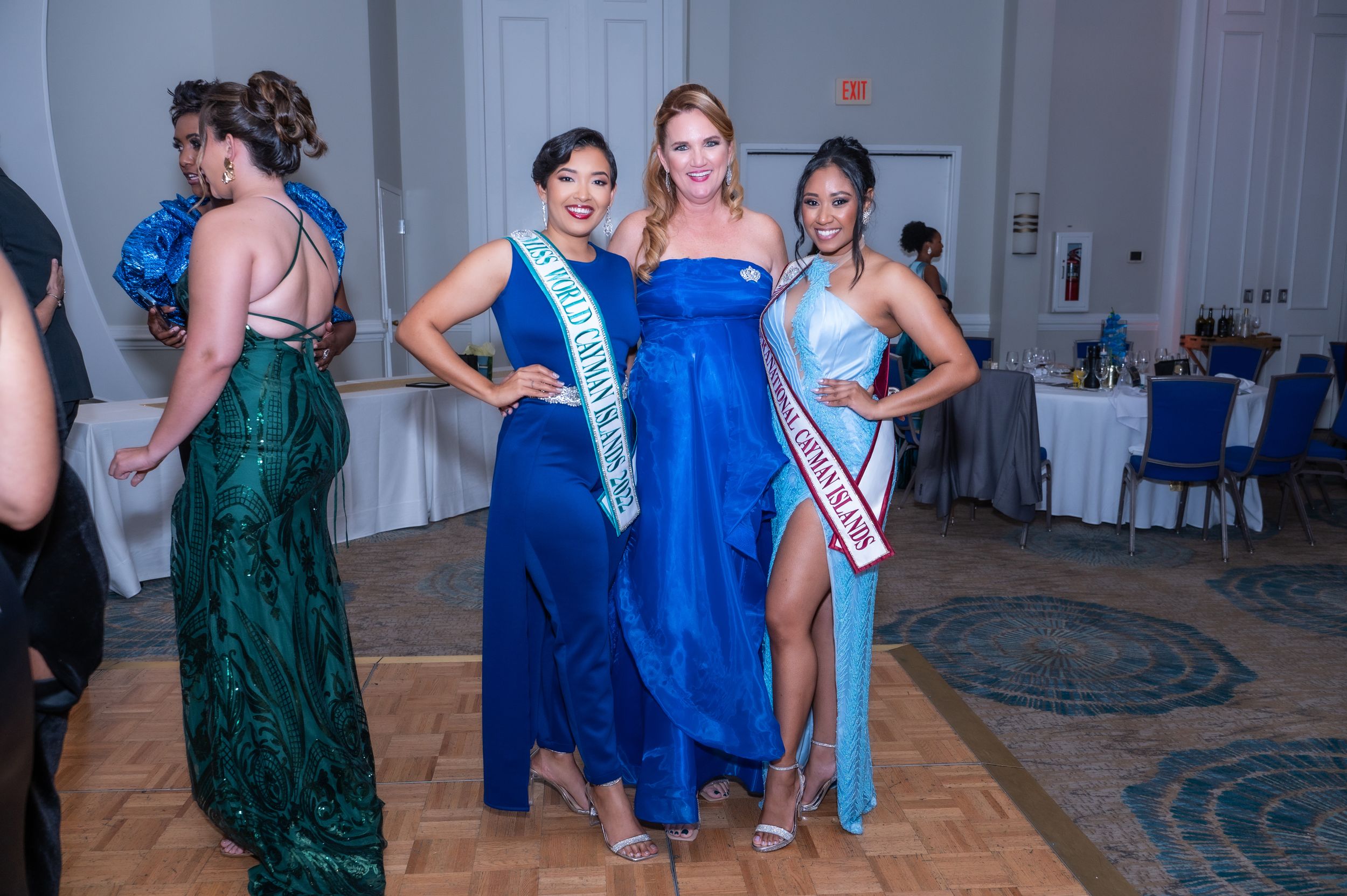 Three women in elegant blue gowns wearing pageant sashes pose together at a formal pageant event.