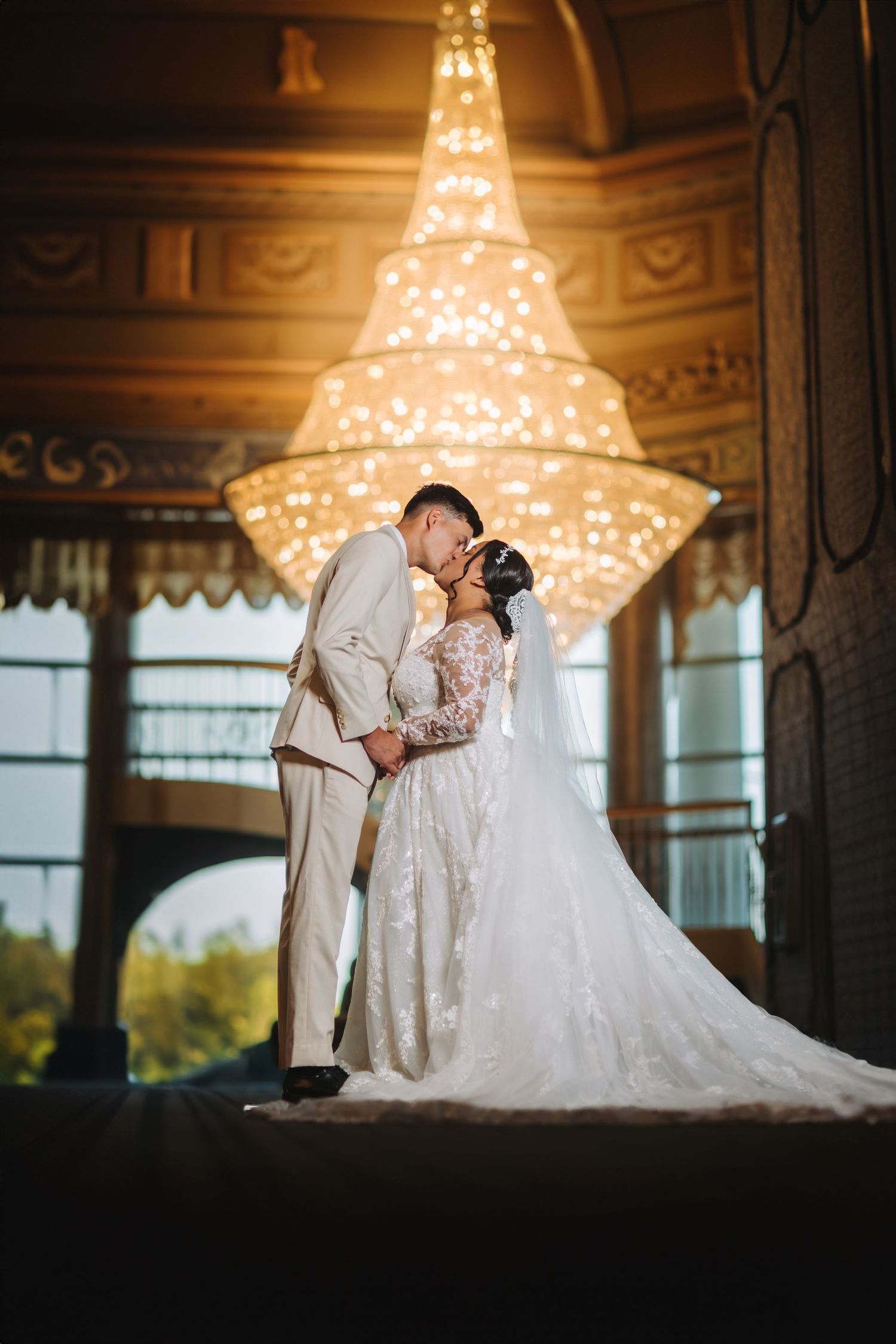 Romantic wedding couple sharing a kiss beneath an ornate crystal chandelier in an elegant ballroom venue.