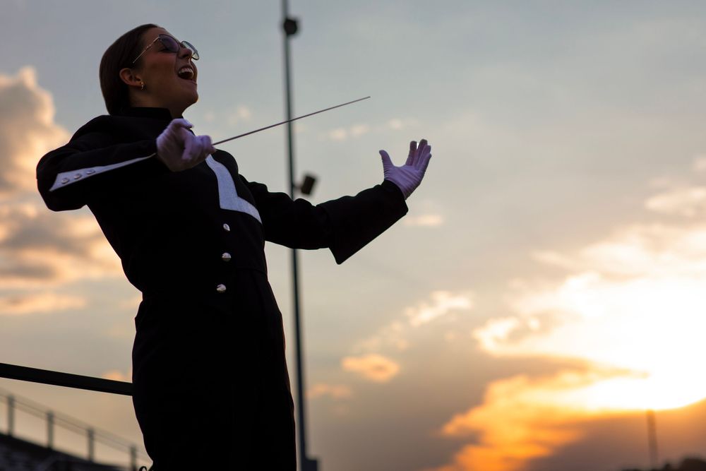 Band conductor in formal attire directing with expressive gestures at sunset.