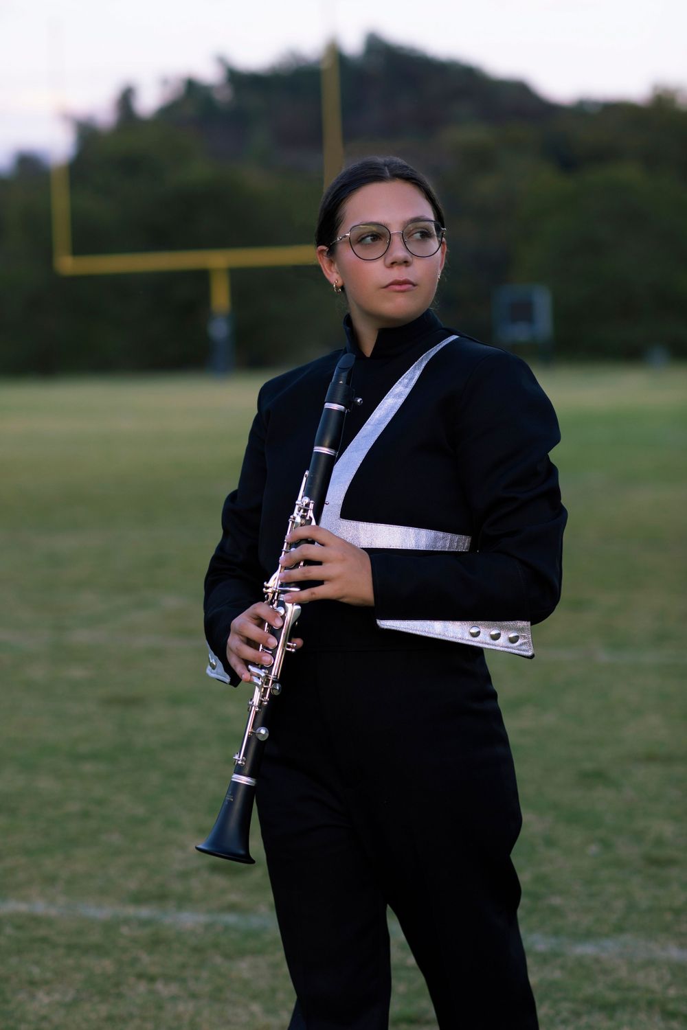 Marching band member in black uniform holding clarinet on football field with goal post in background.