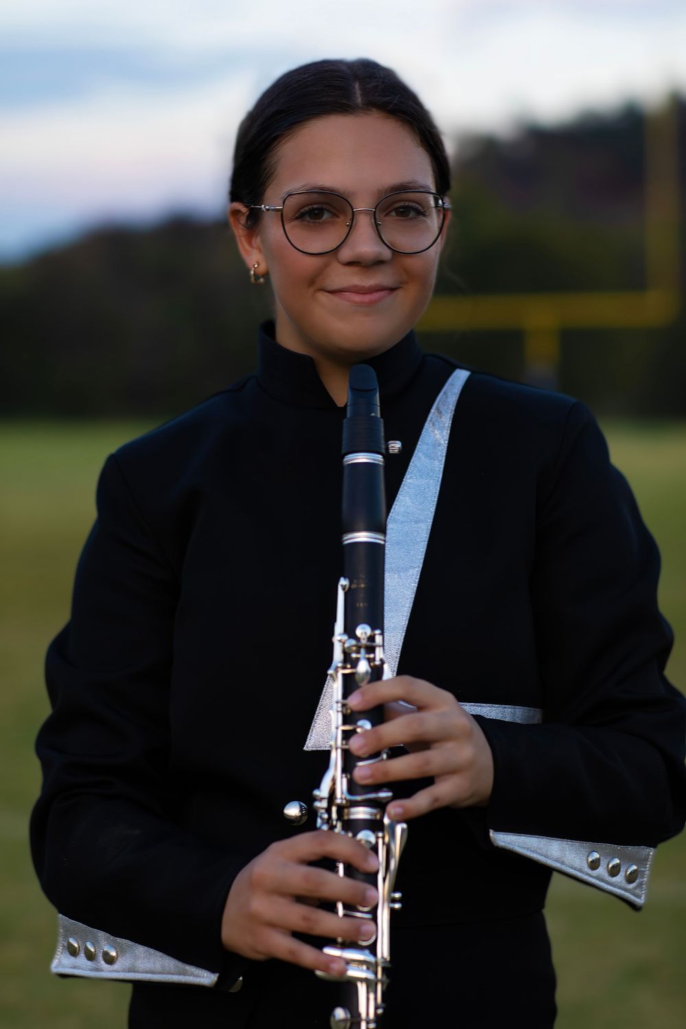 A musician in formal attire holds a clarinet on a football field at dusk.