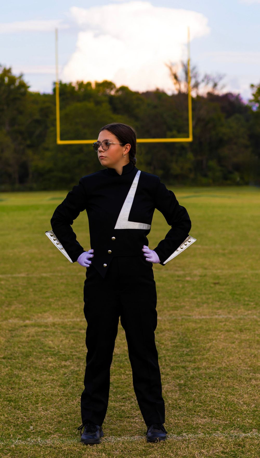 Band member in formal uniform stands confidently on field with football goal post visible.