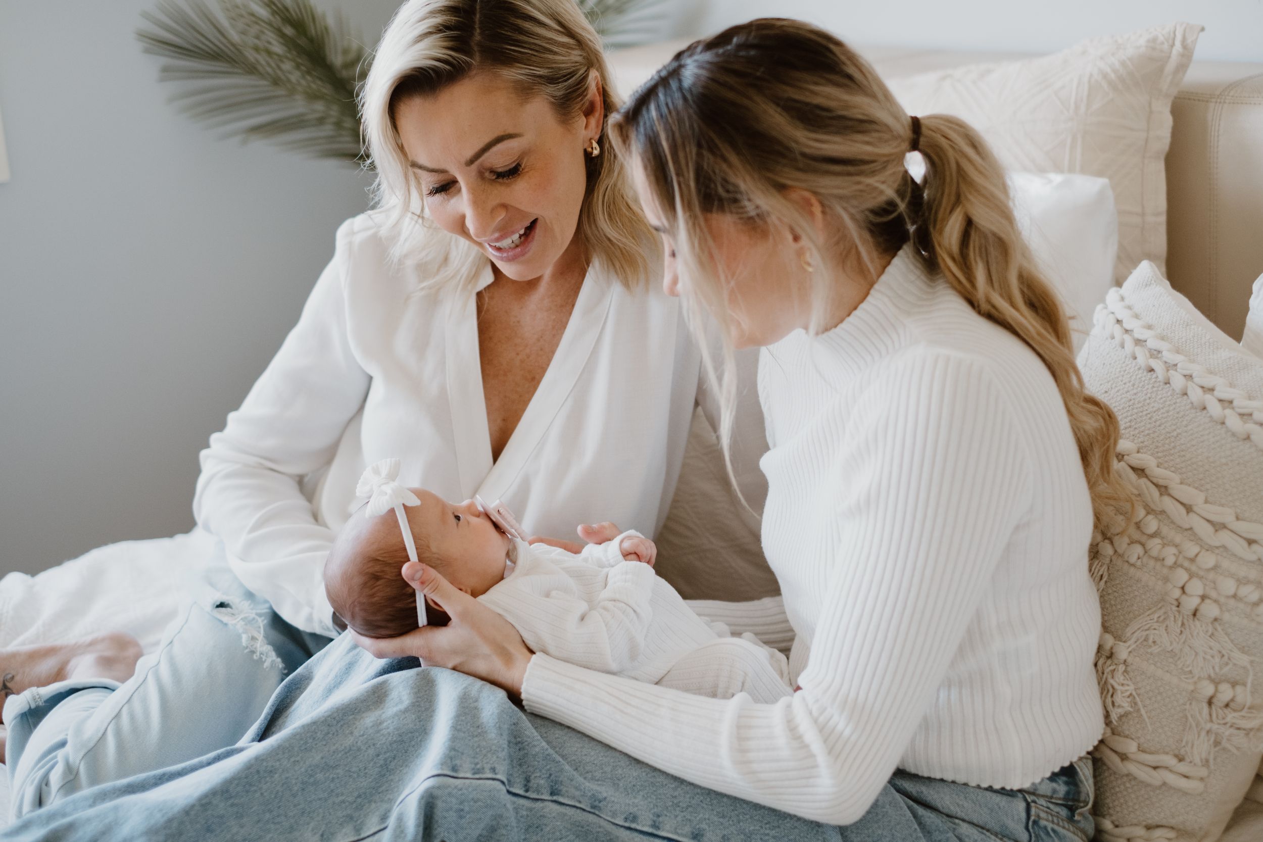 Two women in white clothing sit together on a couch sharing a tender moment with a small infant.