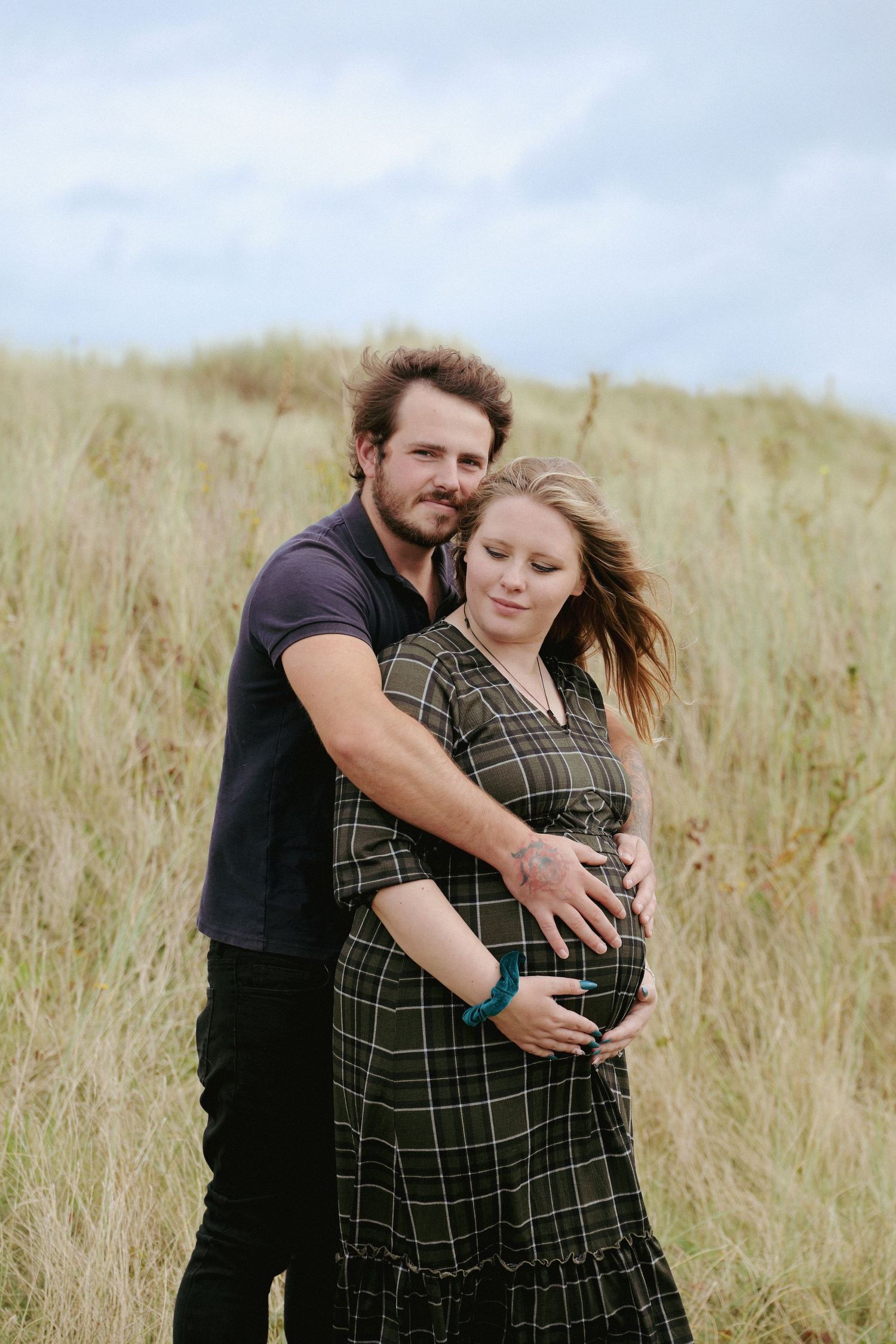 A tender maternity moment captured in tall grass with a couple embracing and cradling a baby bump.