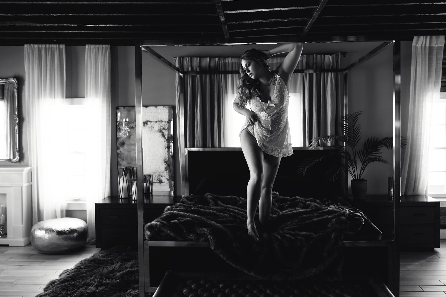 Black and white boudoir photograph showing a silhouette figure standing on a bed in a softly lit bedroom.