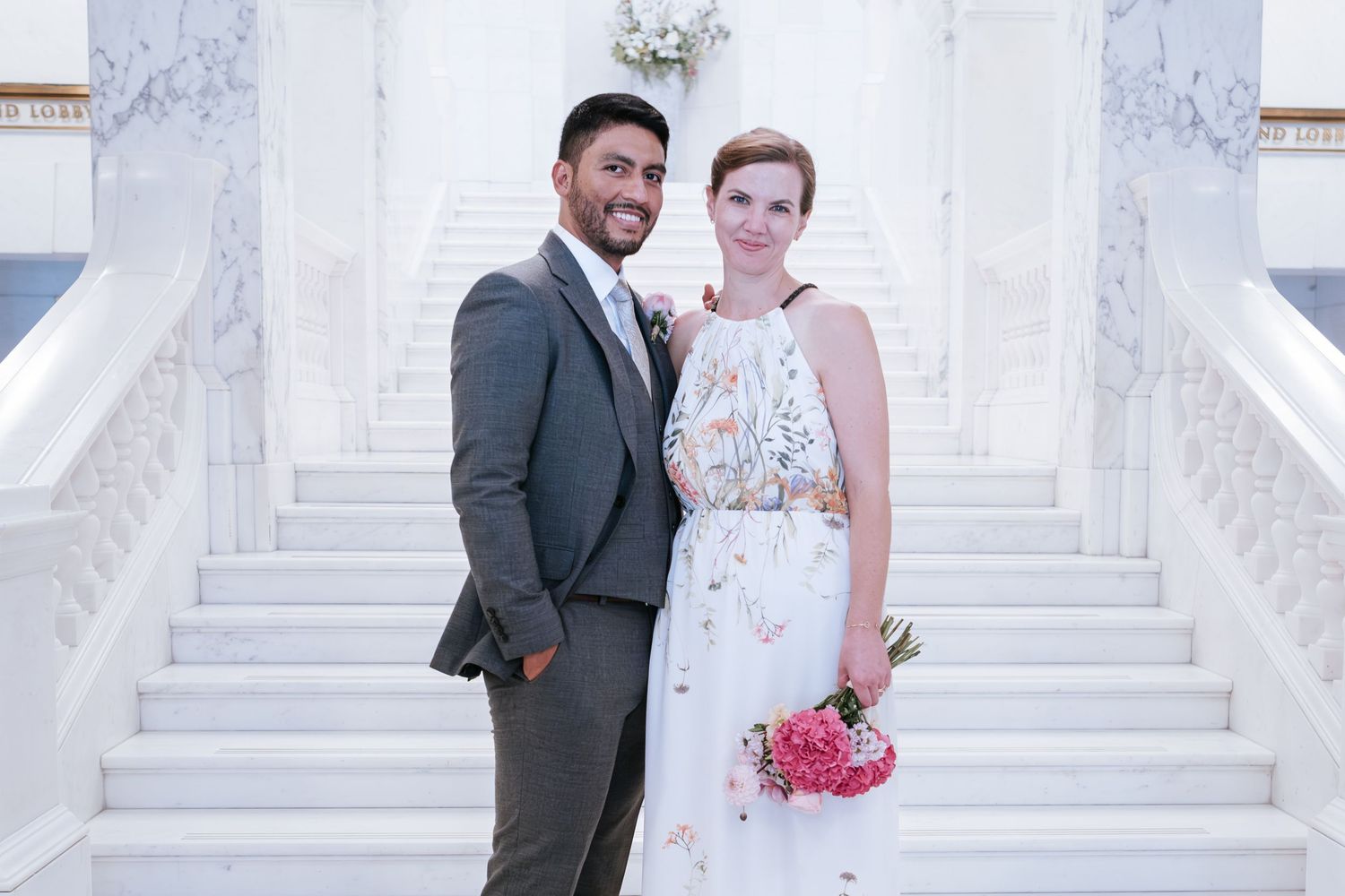 Smiling bride and groom posing on elegant white marble staircase, bride holding pink bouquet in floral wedding dress.