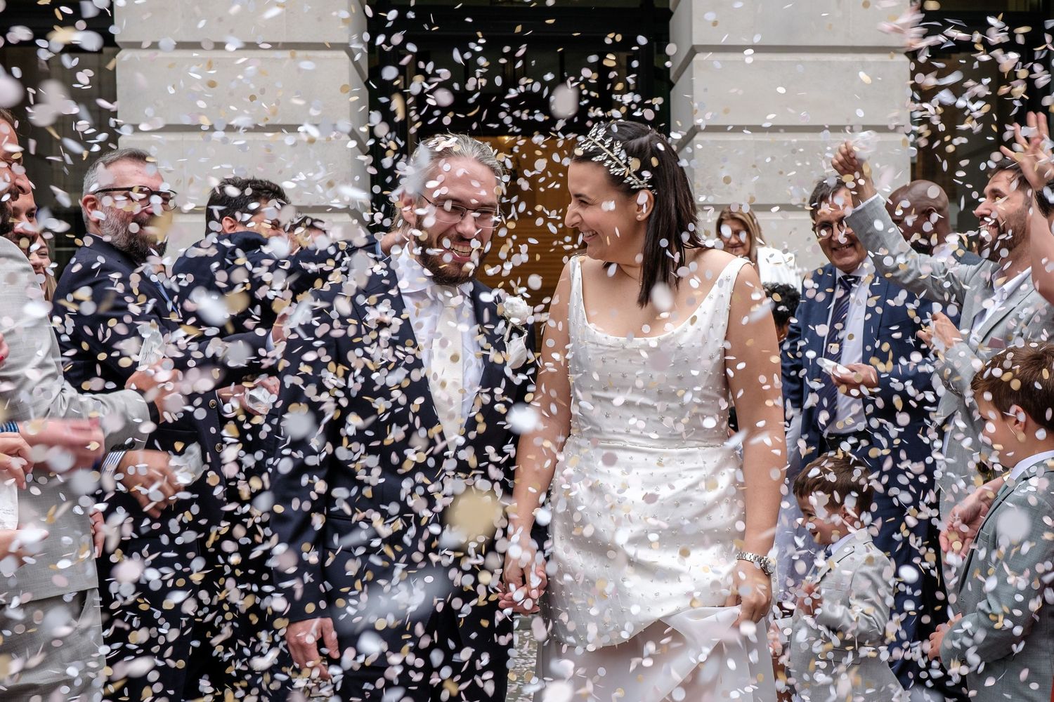 Bride and groom walk through confetti shower surrounded by wedding guests outside a building.