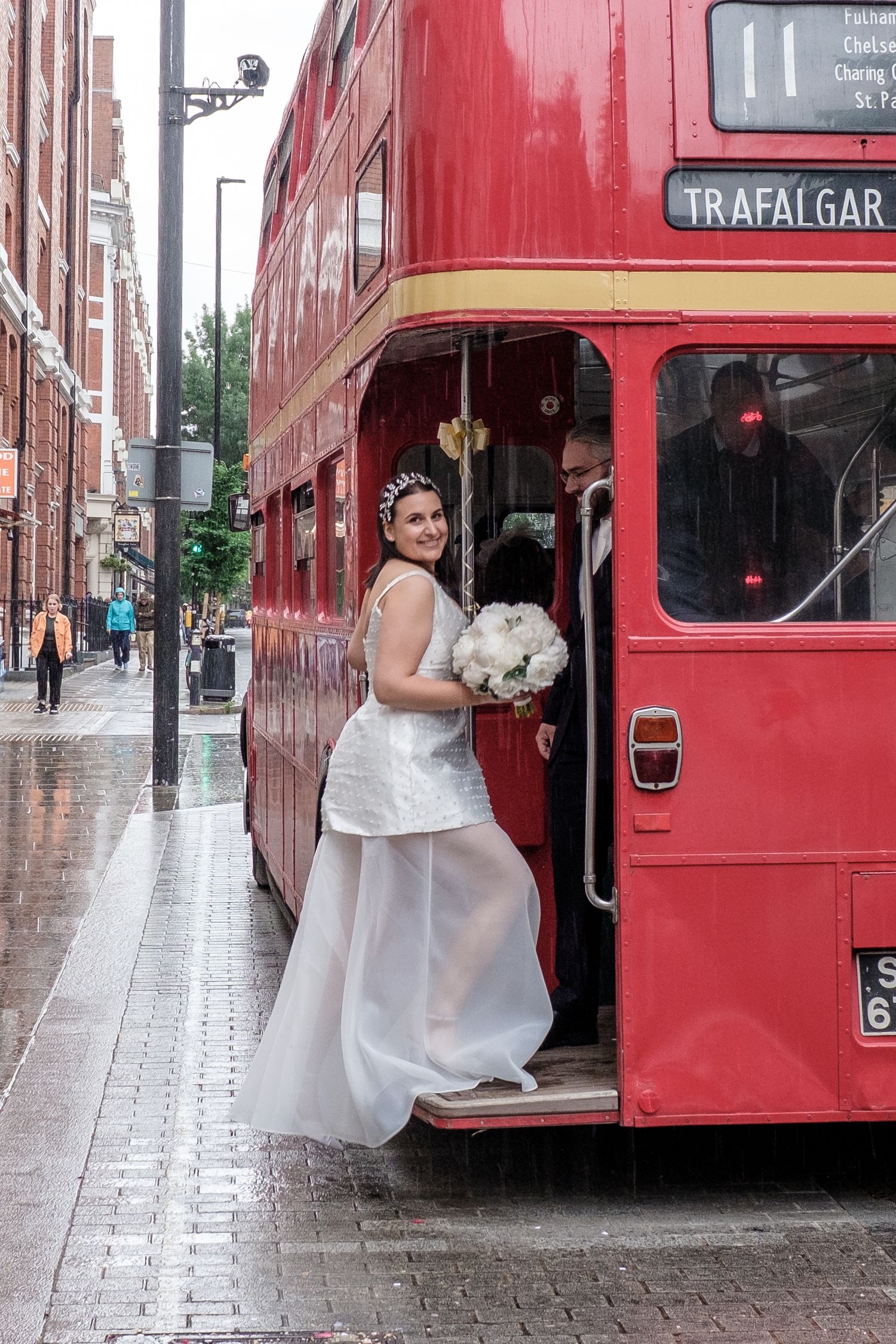 Bride in white gown holding bouquet boards red double-decker London bus to Trafalgar Square on a rainy wedding day.