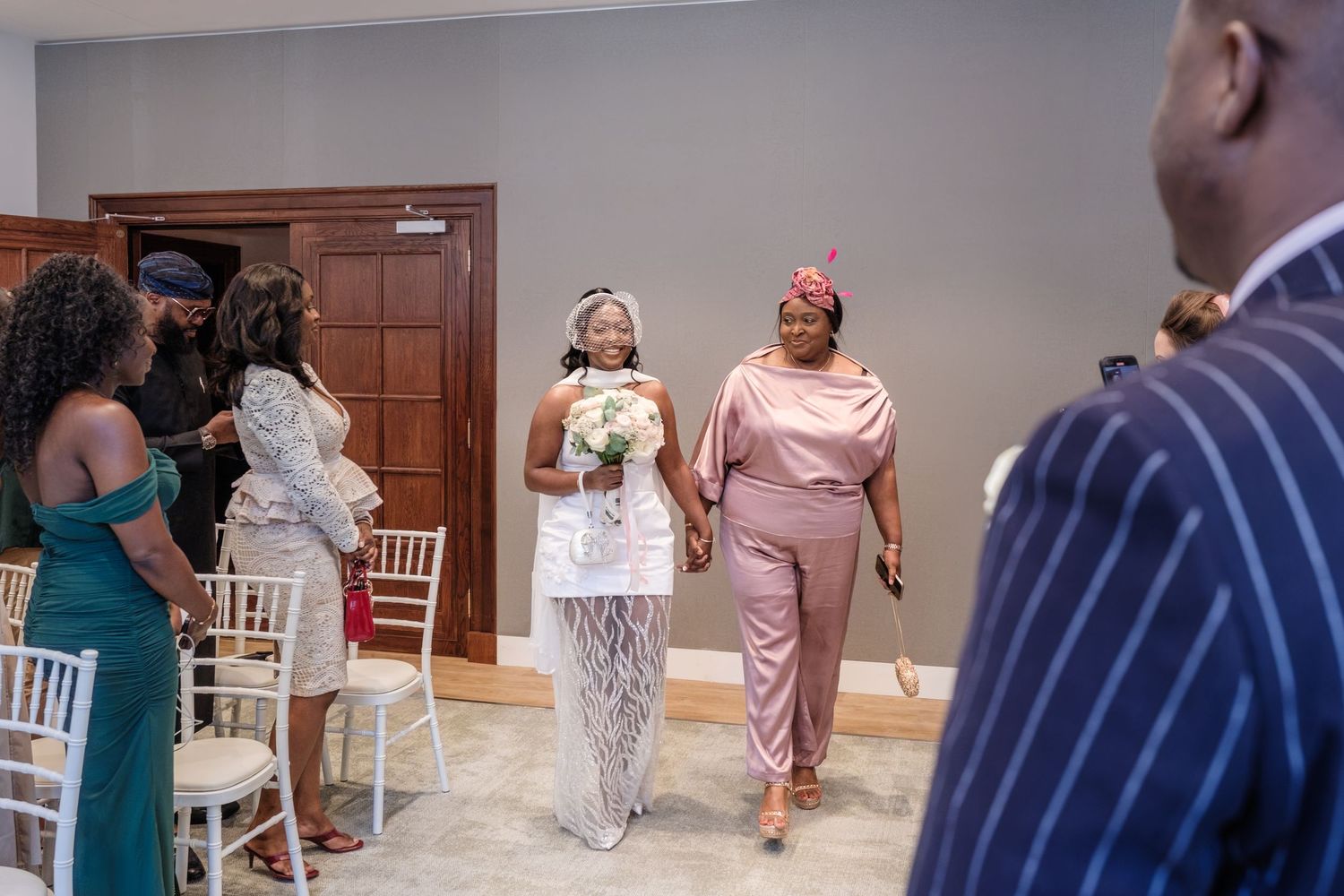 Bride walking down the aisle with escort at an elegant indoor wedding ceremony with white chairs.