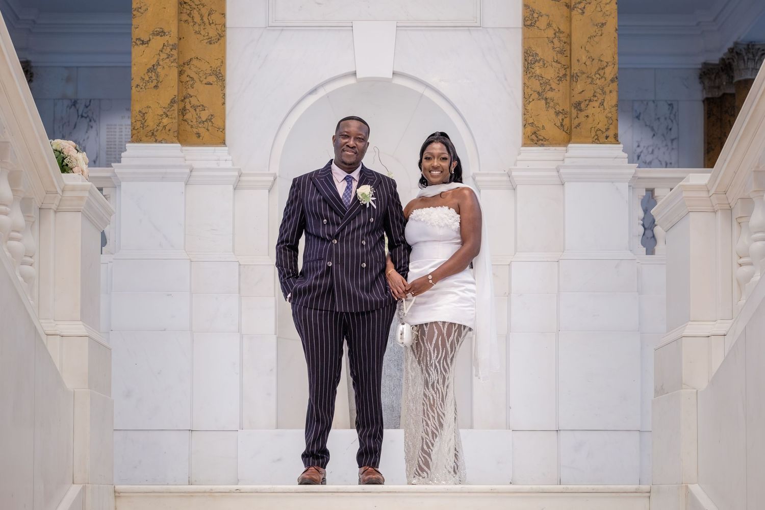 Elegant couple posing on white marble stairs in wedding attire, groom in pinstripe suit, bride in white mini dress.