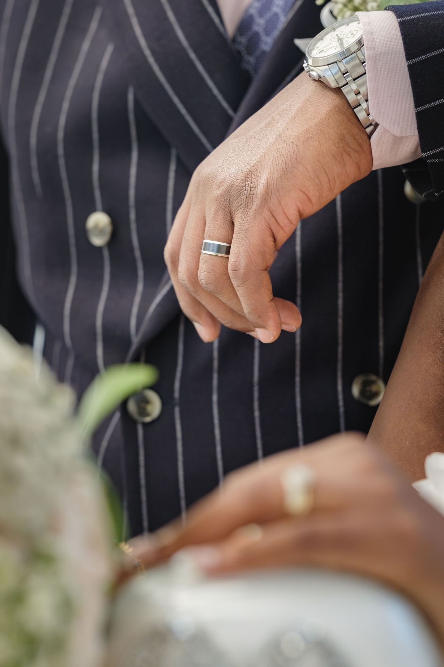 Close-up of groom's hand with black wedding band and silver watch, beside bride's hand with gold ring.