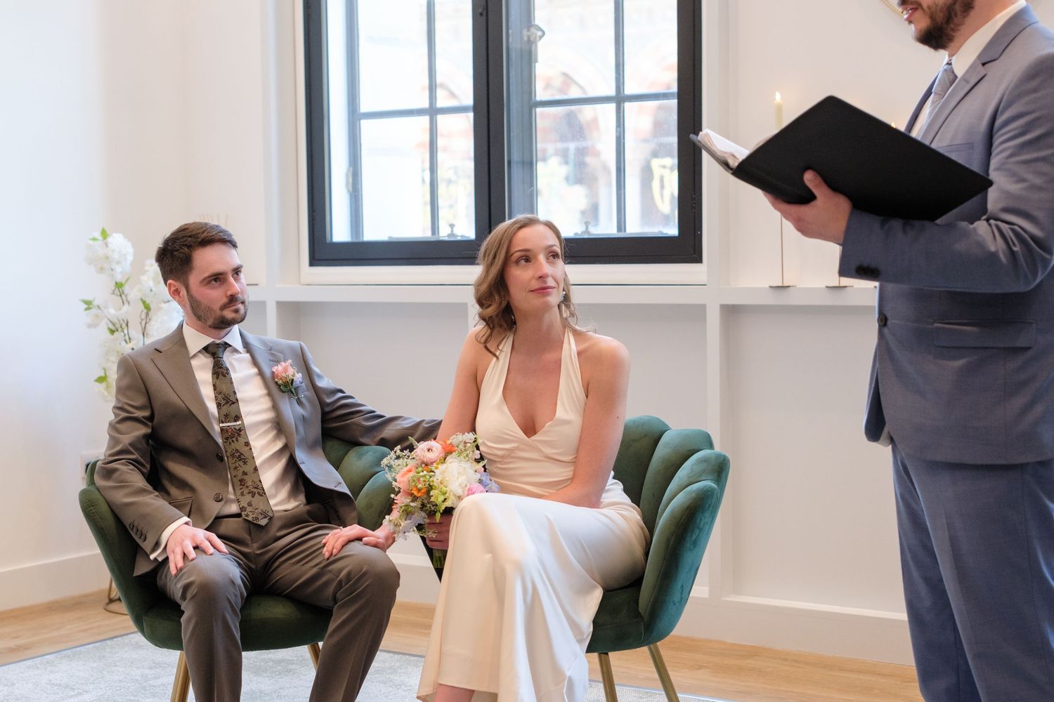 Bride and groom seated at civil wedding ceremony while officiant reads from black binder in bright white room.
