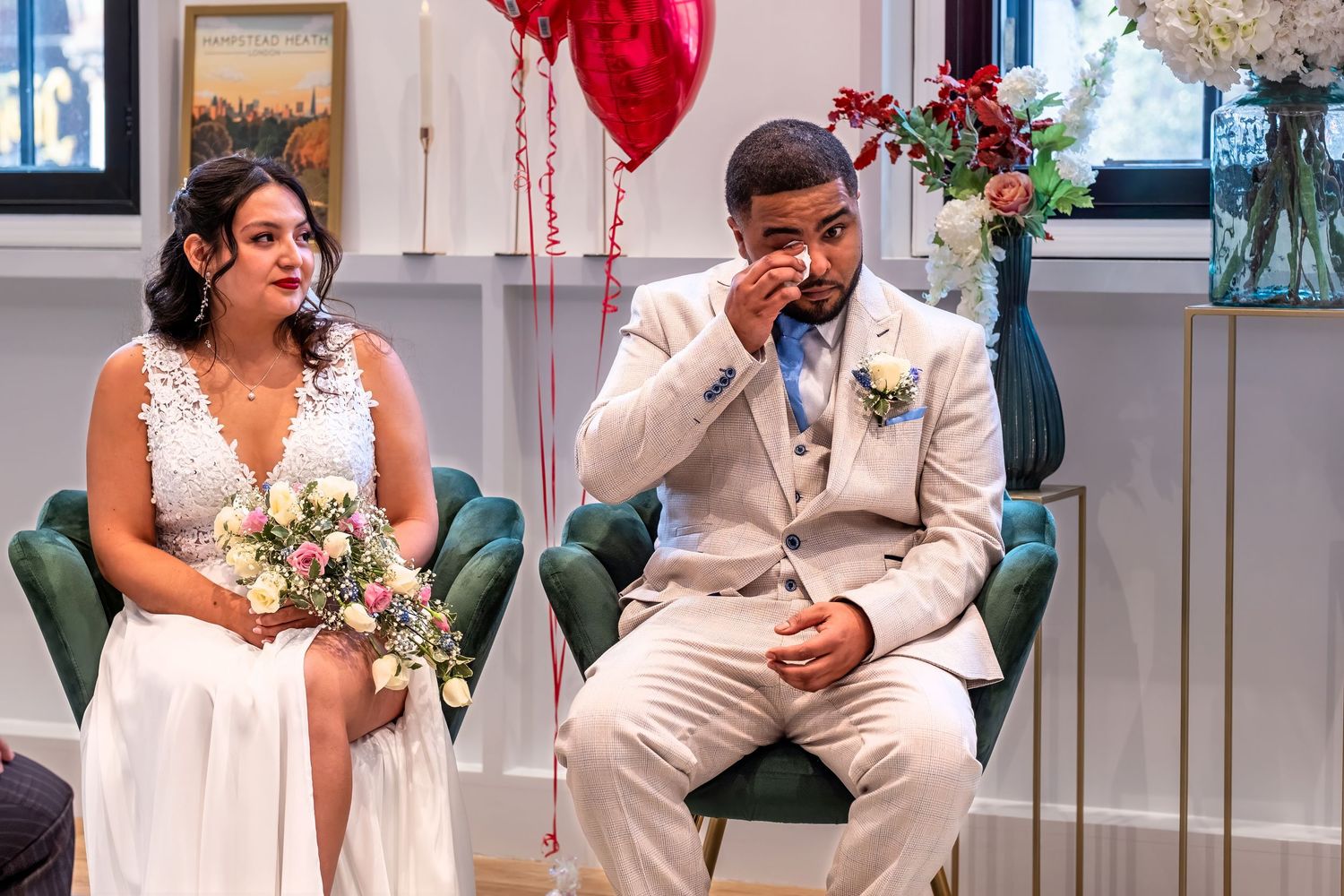 Emotional groom wipes tears beside bride holding bouquet at wedding ceremony with red heart balloons.
