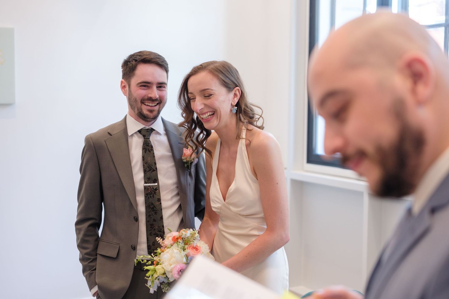 A smiling bride holding flowers and groom in a gray suit laugh joyfully during their intimate wedding ceremony indoors.