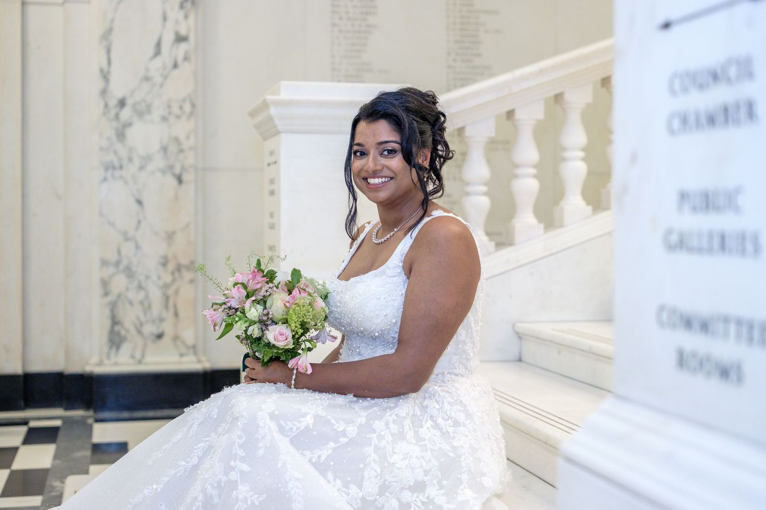 Radiant bride in lace ball gown holding pink bouquet, seated on grand staircase inside ornate building.