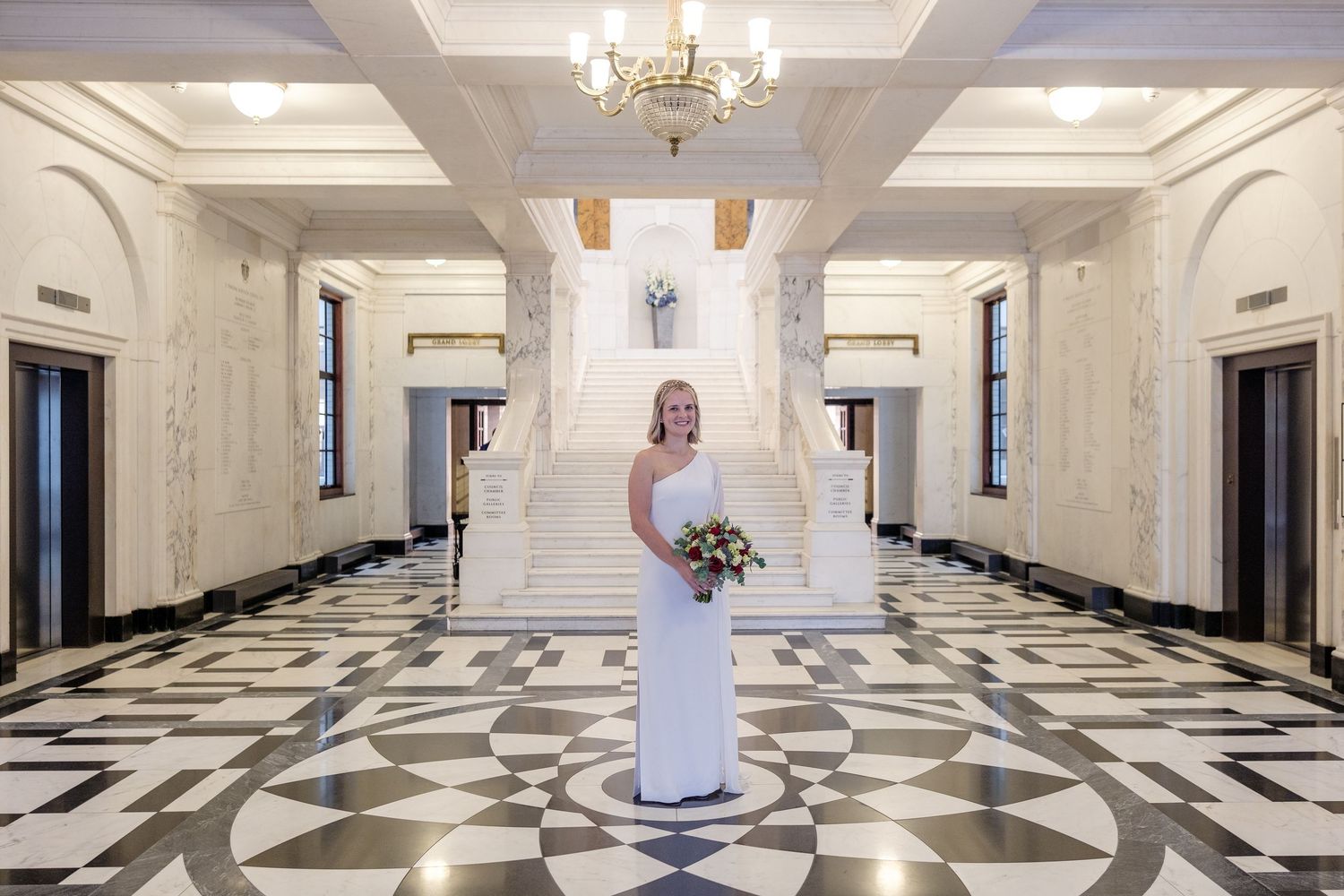Bride in white gown poses in elegant marble hall with chandelier and grand staircase, holding a bouquet.