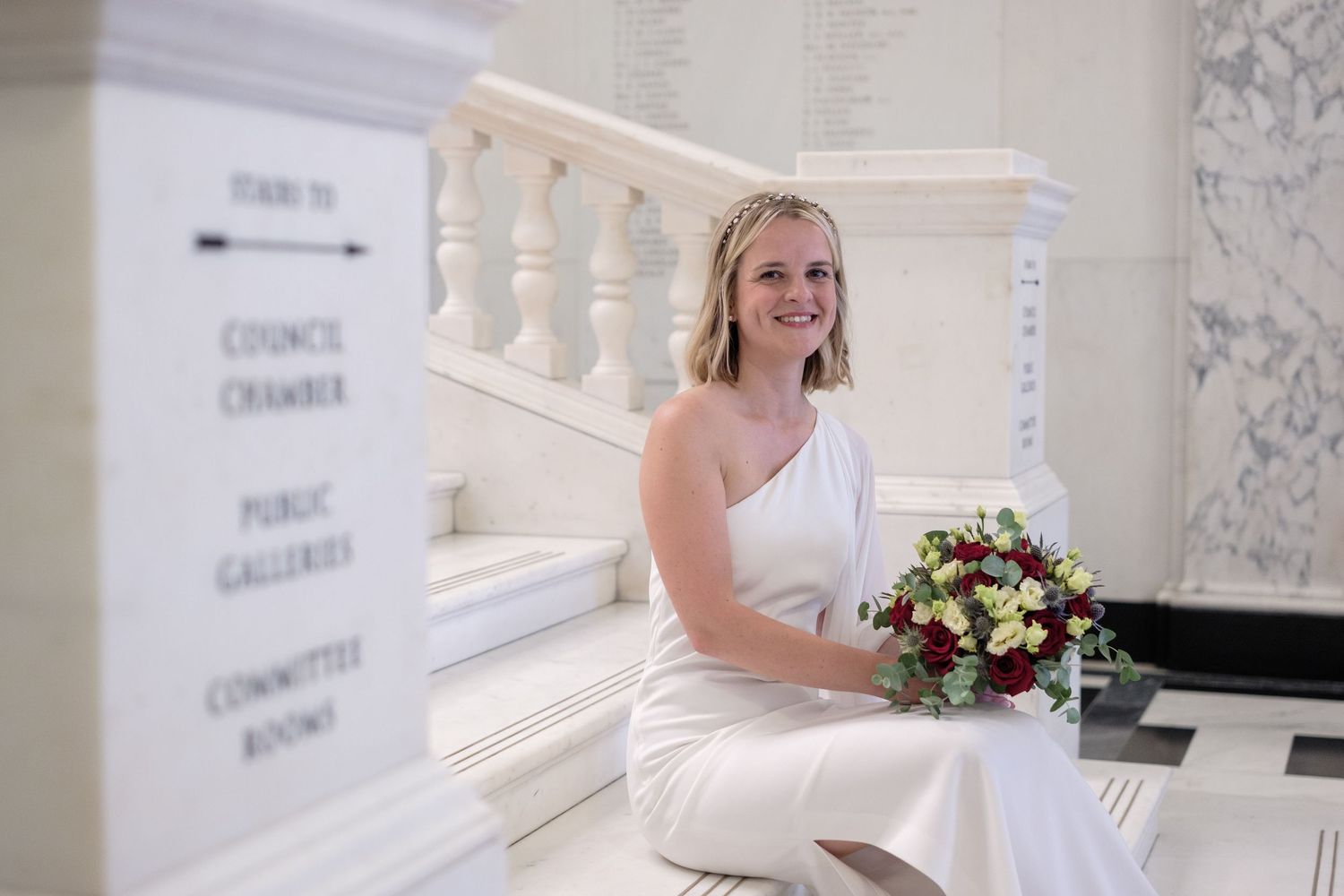 Bride in white one-shoulder gown sits on marble steps holding red and white bouquet near council chamber signs.