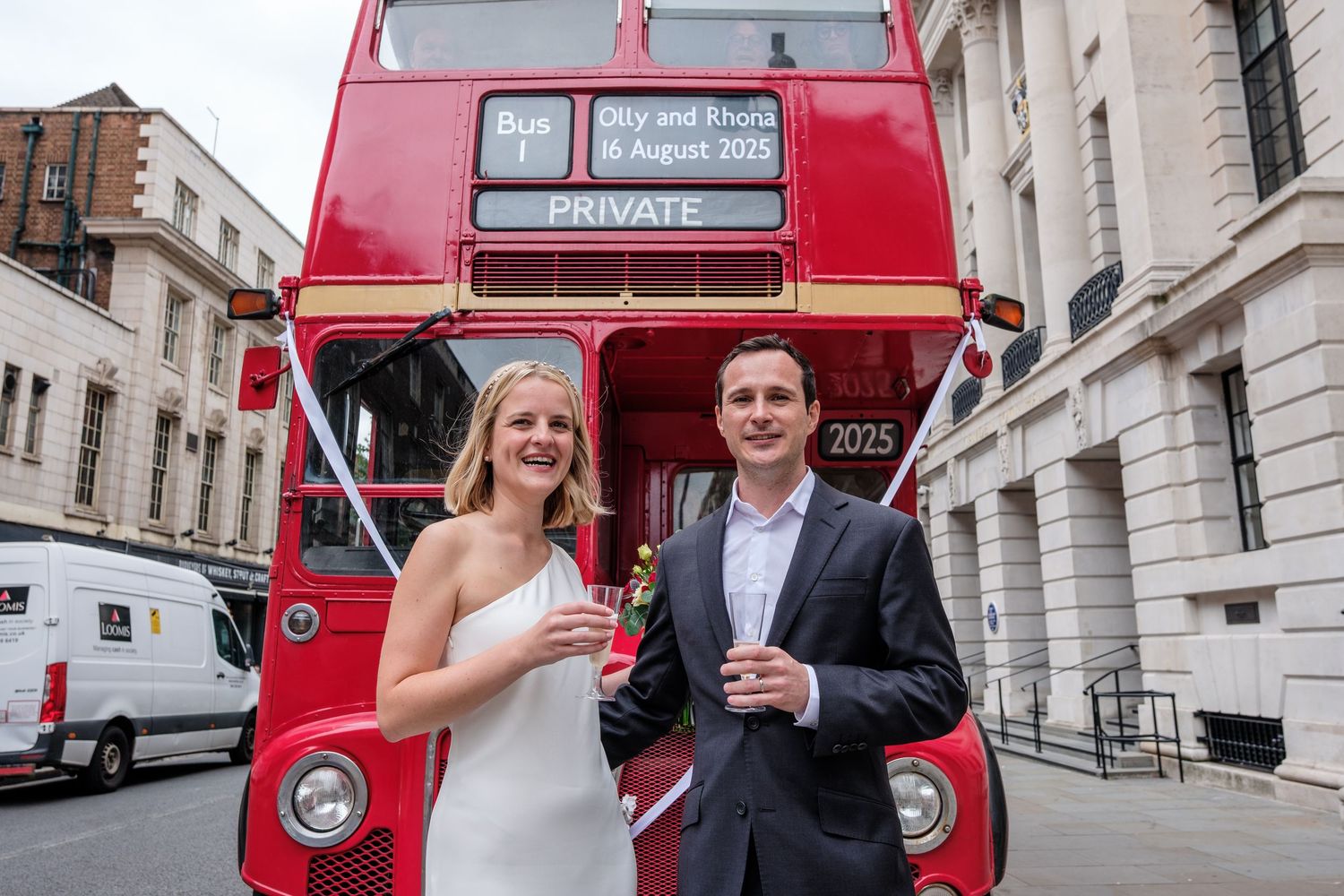 Couple celebrating in front of a red double-decker private bus in London, holding champagne glasses.