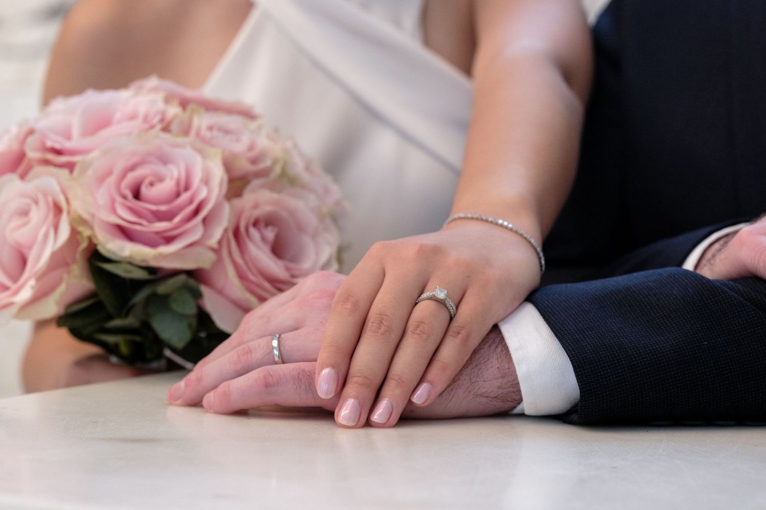Newlywed couple holding hands with wedding rings, bride holding pink roses bouquet on white table.