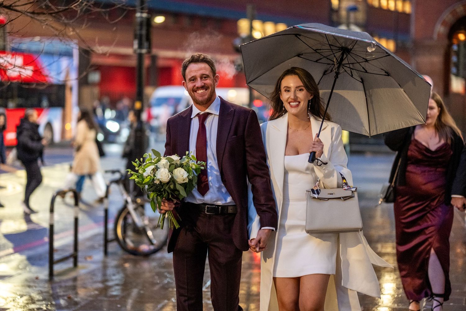 Couple in wedding attire walking in rainy city street, man holding flowers, woman with umbrella.