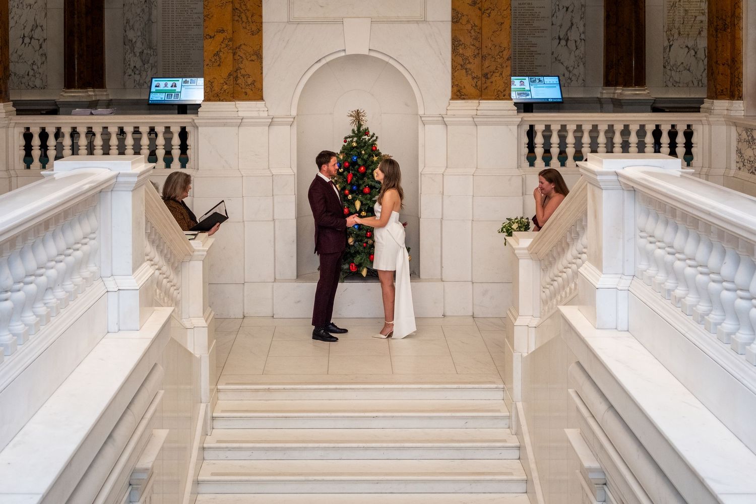 Couple exchanging vows on grand marble staircase with Christmas tree in elegant white building interior.