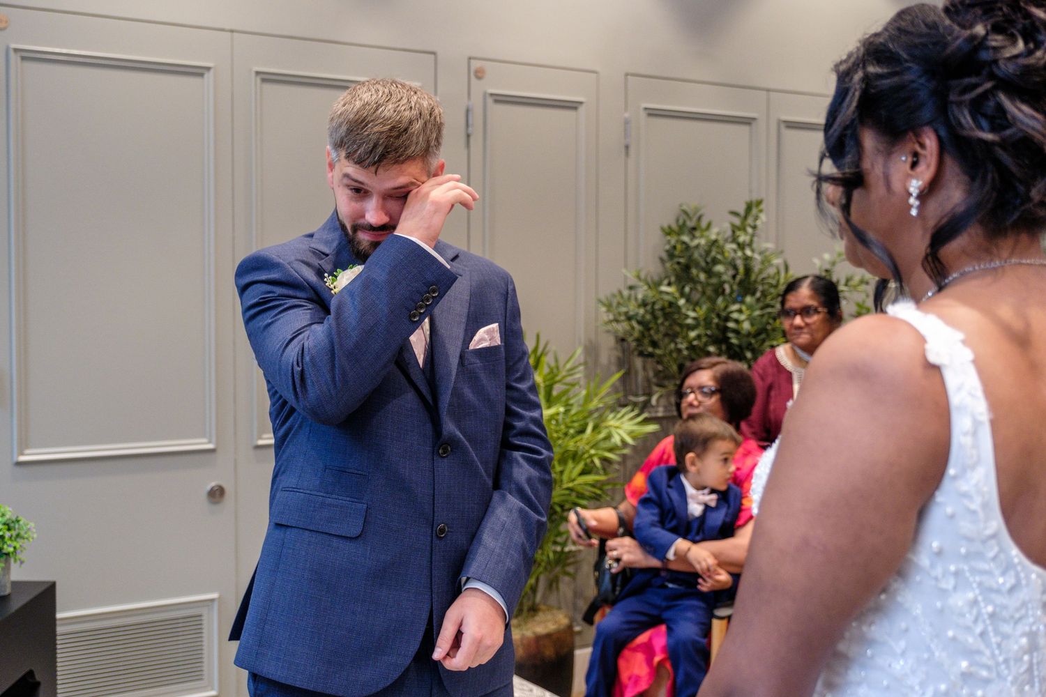 Emotional groom wiping tears during wedding ceremony while bride stands nearby with guests seated in background.