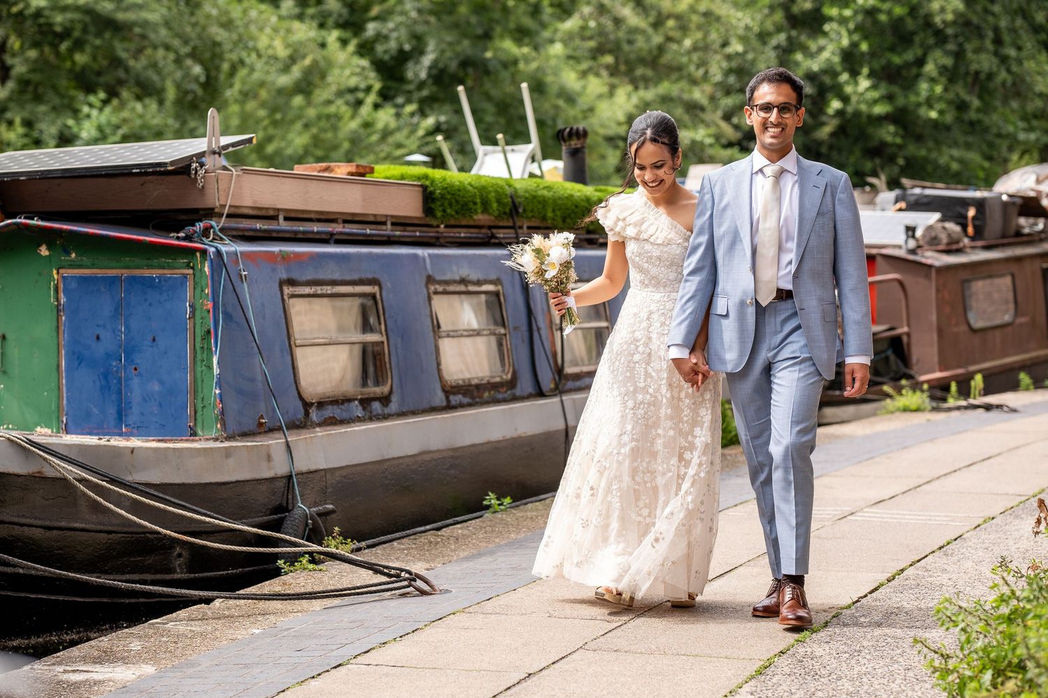 Bride and groom walking hand in hand along a canal towpath with colorful narrowboats in the background.