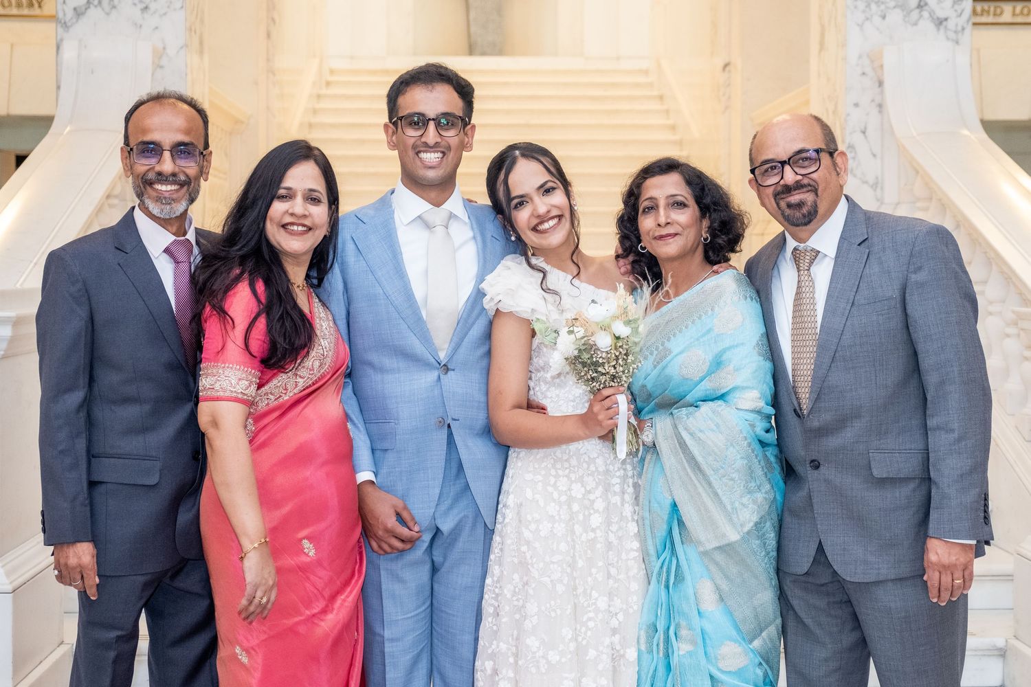 A joyful South Asian family poses together at a wedding, with the bride in white and guests in colorful traditional attire.