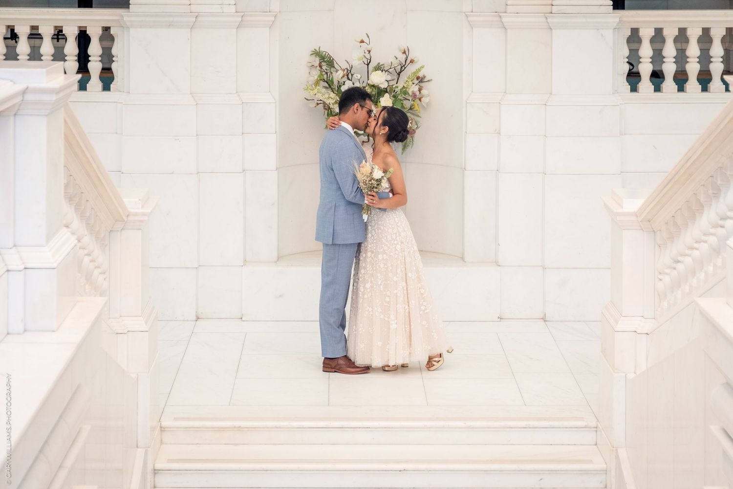 Bride and groom sharing a romantic kiss on white marble steps with floral arch backdrop at elegant wedding venue.