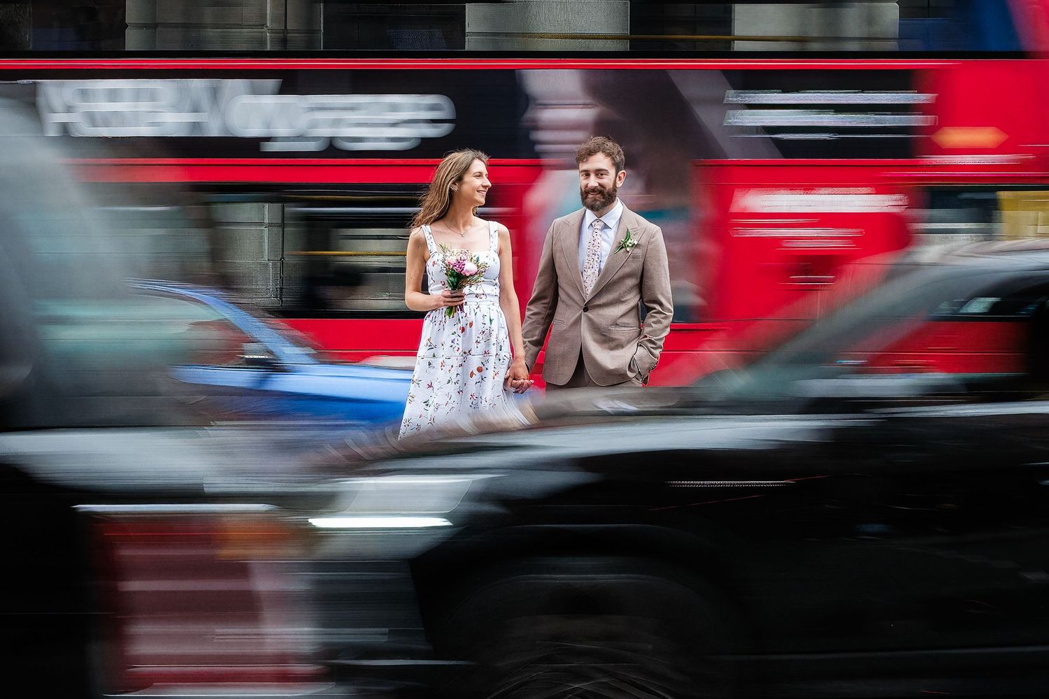 Couple in formal attire walking through busy London street with red double-decker bus and black taxi cab in motion blur.