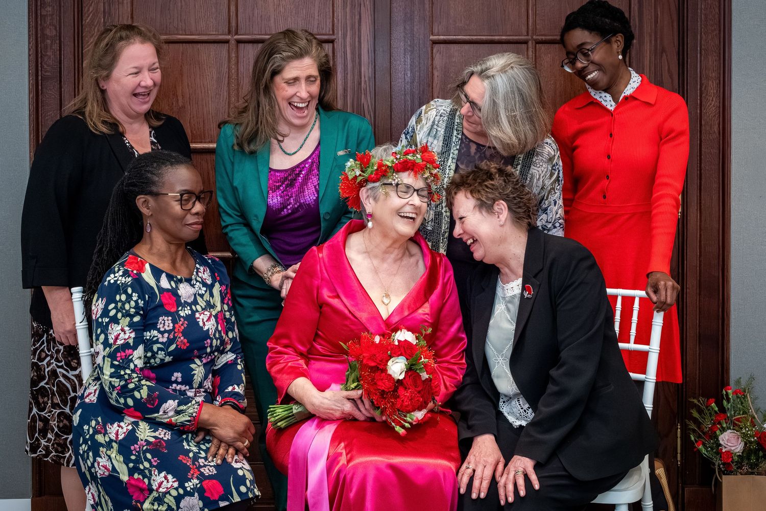 Group of seven women posing together, smiling and laughing, with one woman in red dress holding bouquet.