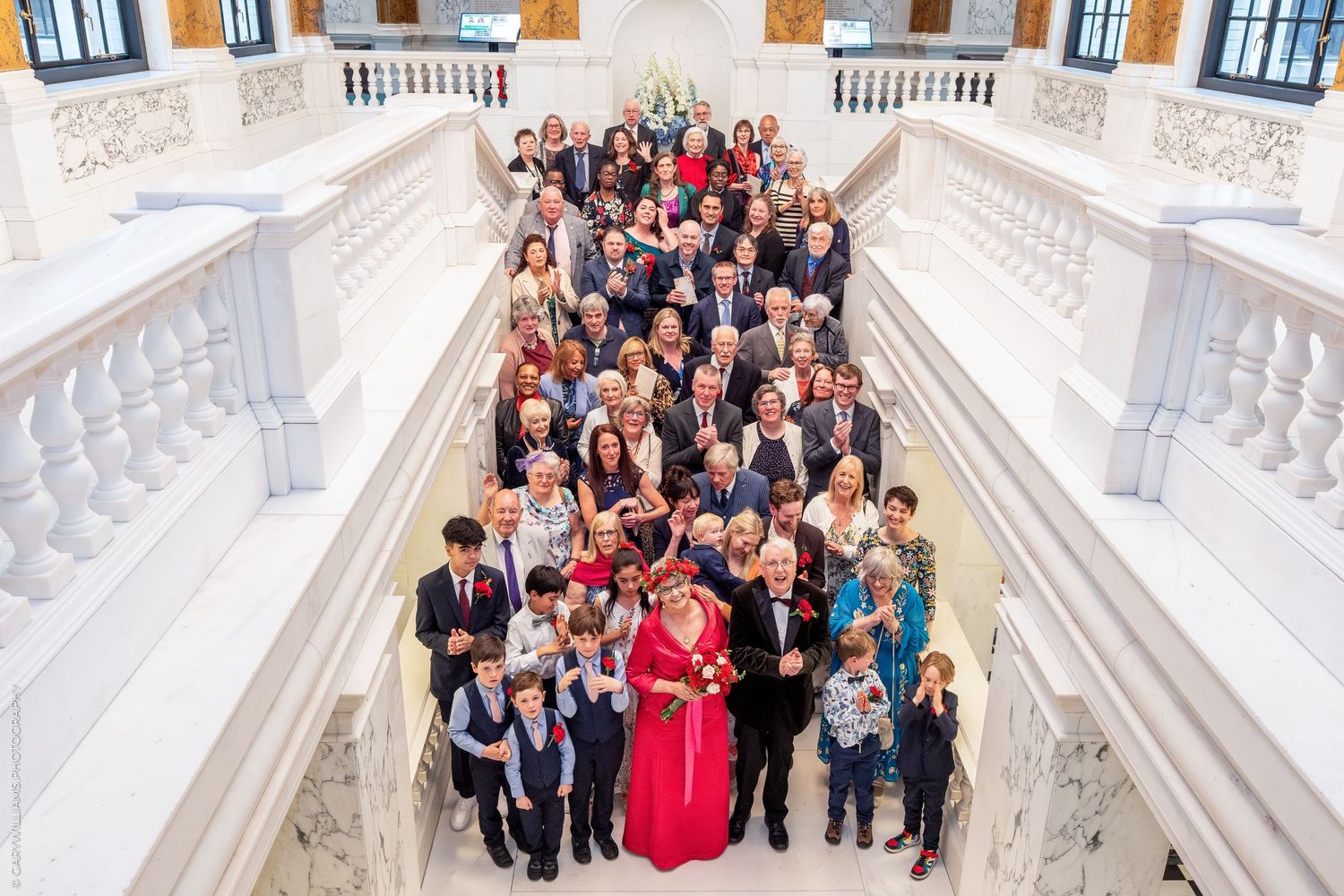 Large wedding group photo on grand white marble staircase with bride in red dress surrounded by family and guests.