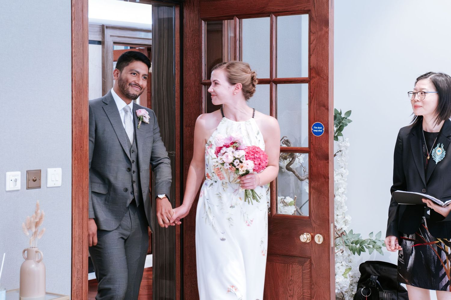 Smiling interracial couple holding hands in wedding attire, bride holding pink bouquet, greeted by officiant indoors.