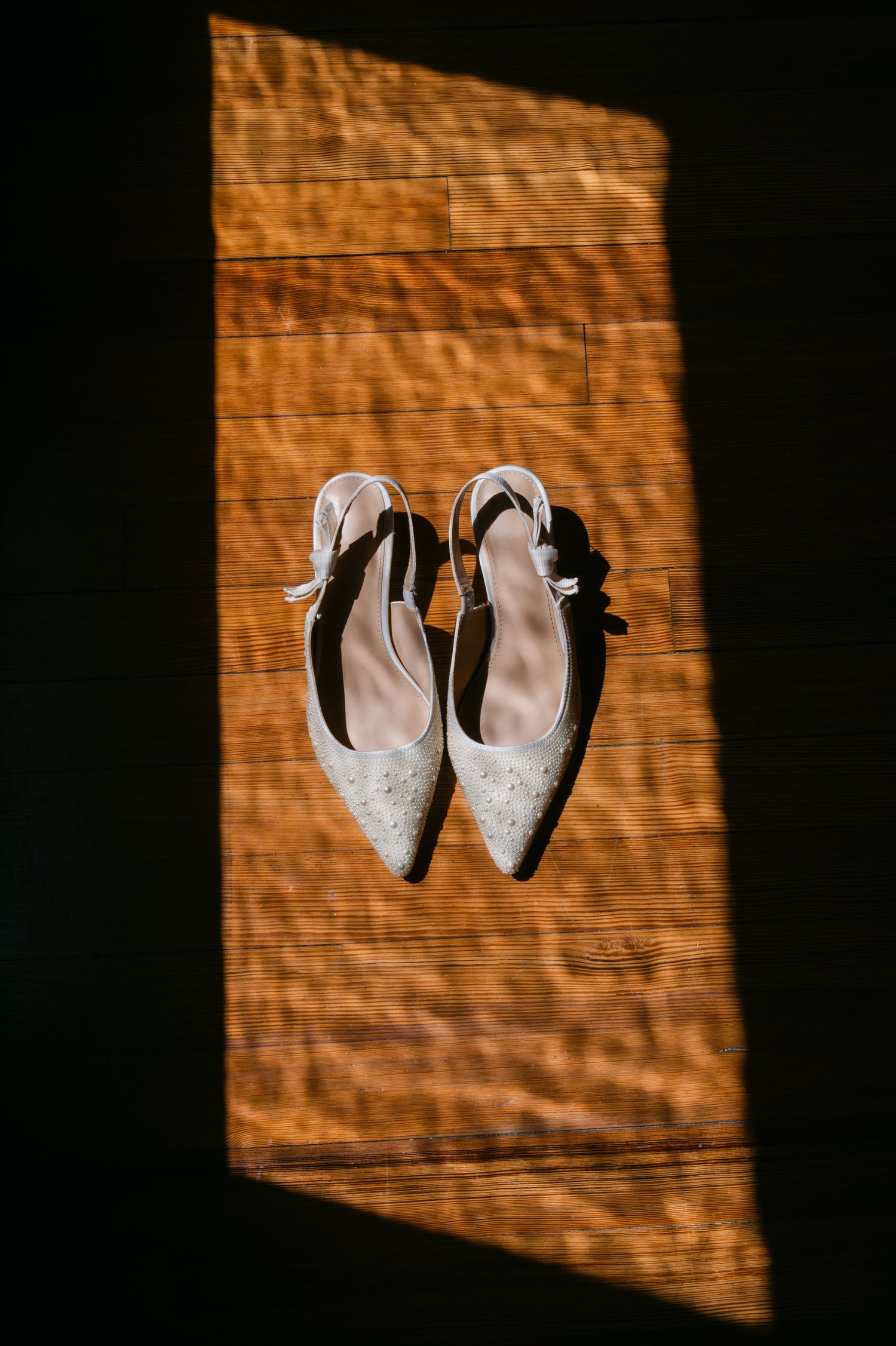 Elegant white bridal heels with ankle straps displayed on a wooden floor in a beam of natural sunlight.