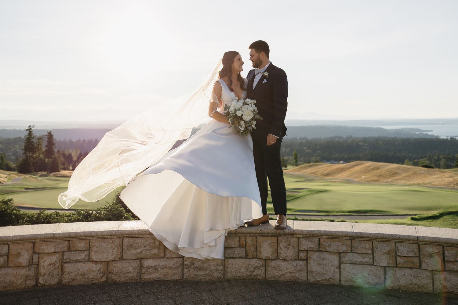 Bride and groom share a romantic moment on a stone wall overlooking a golf course, with veil flowing in the wind.