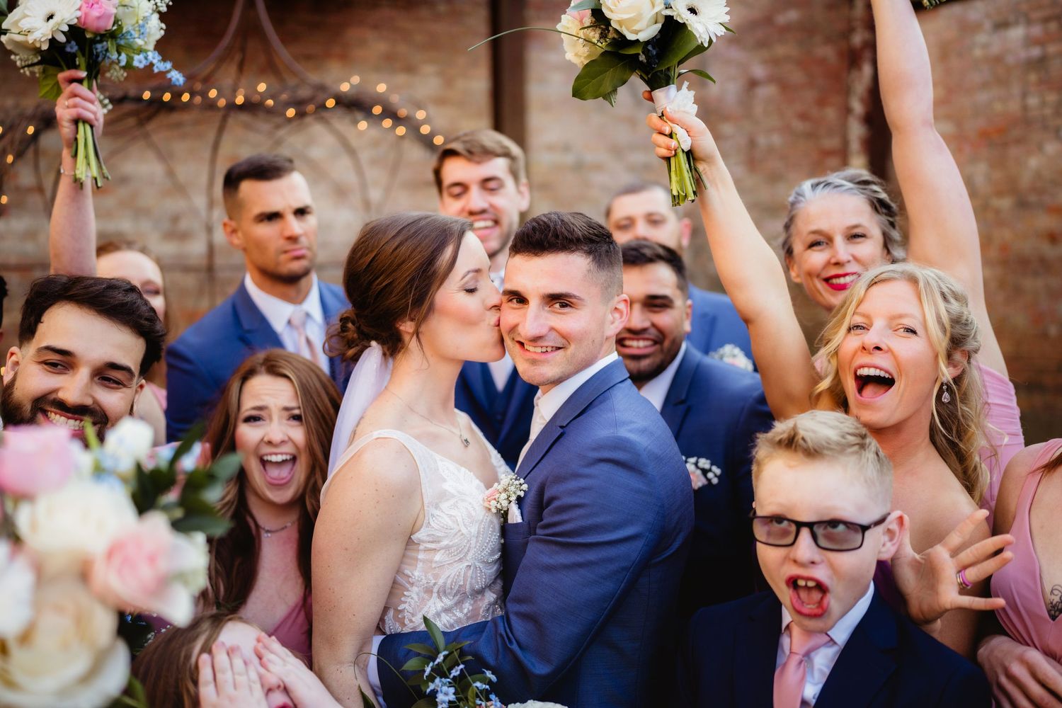 Joyful wedding couple kissing surrounded by cheering bridal party holding bouquets in a rustic brick venue.