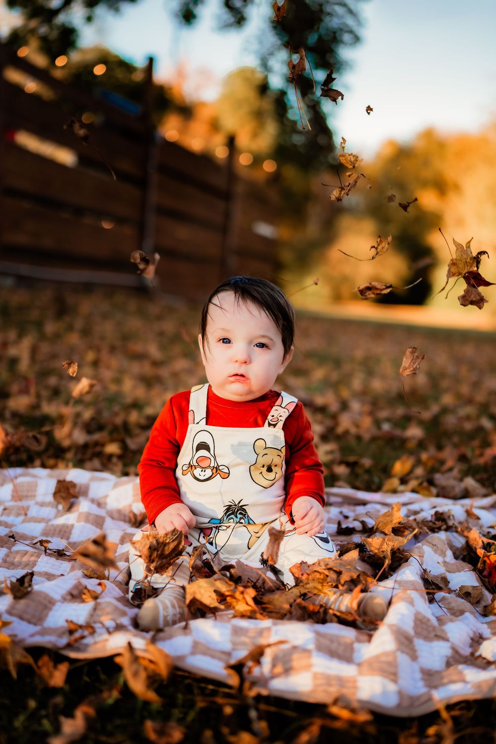 Baby in Winnie the Pooh overalls sits on a quilt surrounded by autumn leaves in a sunny backyard.
