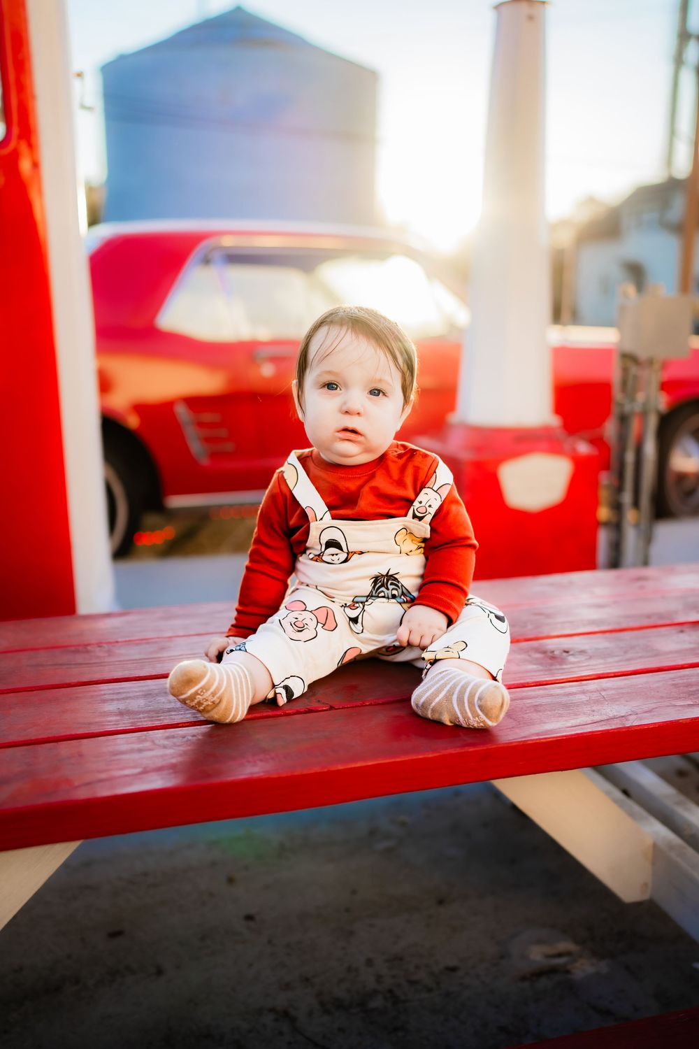 Adorable baby in red top and overalls sitting on a red bench outdoors with a classic red car in the background.