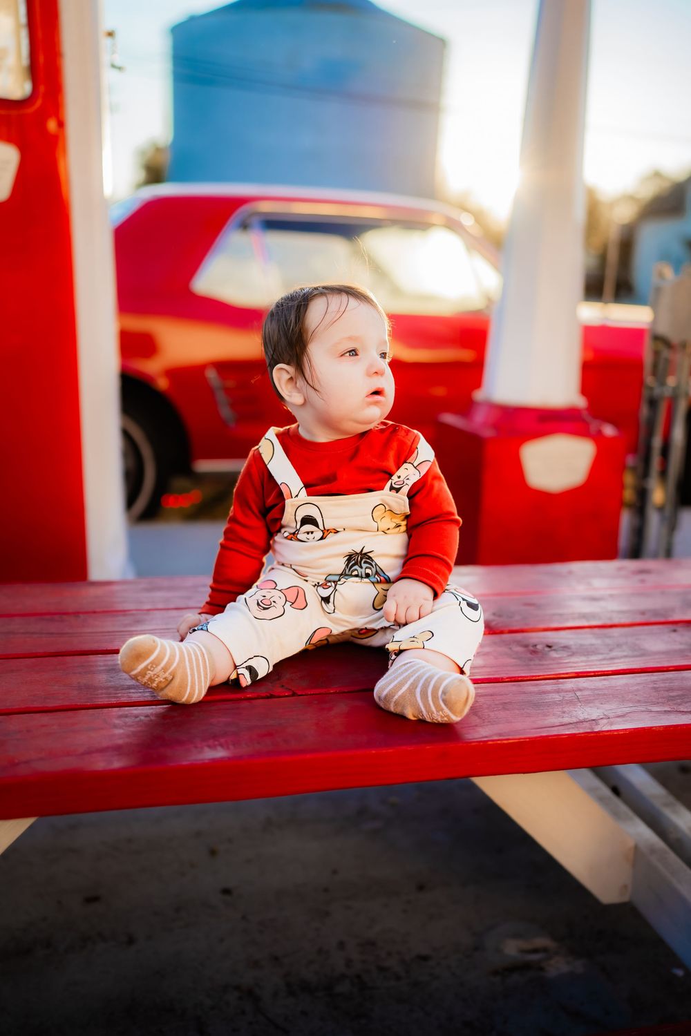 Adorable baby in red shirt and printed overalls sitting on a red bench outdoors near a vintage red car.