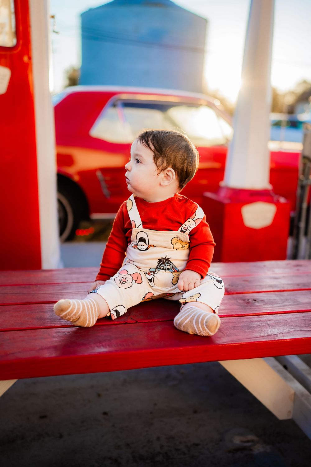 A baby in red shirt and printed overalls sits on a red bench outdoors near a classic red car in golden light.