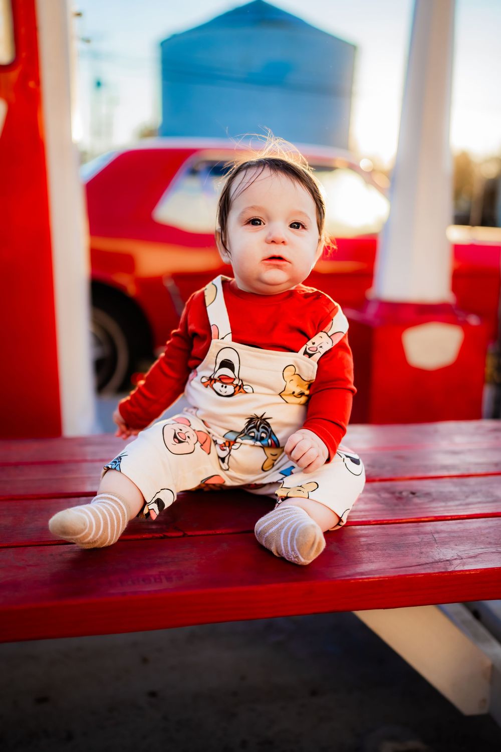 Baby in red shirt and cartoon overalls sitting on a red bench outdoors in warm sunlight.