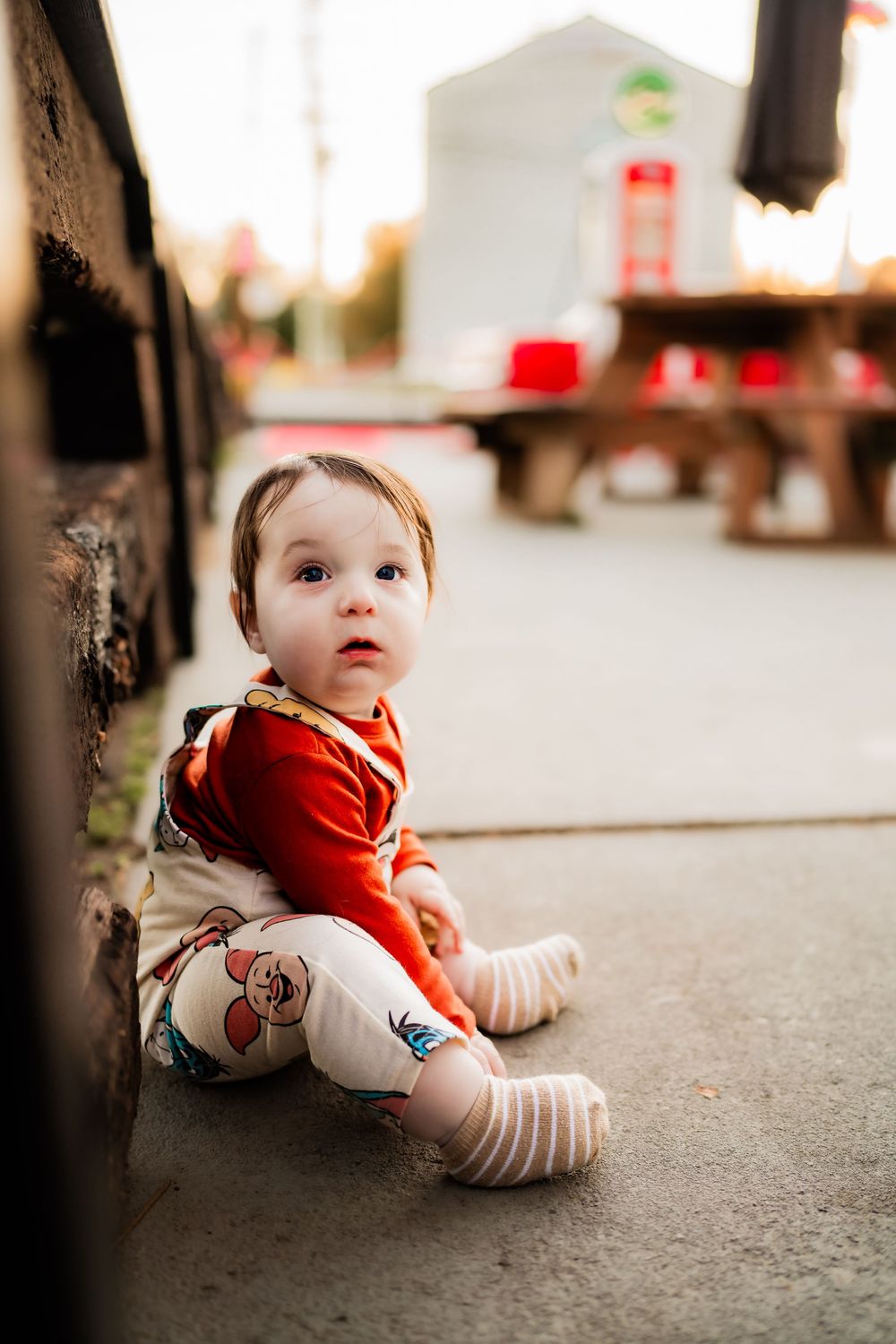 Cute baby in red outfit and striped socks sitting on ground near wooden wall outdoors.